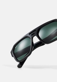 Melni plastmasas saulesbrilles ar stūrainām rāmēm, tumši zaļiem objektīviem un izteiktām sānu izgriezumiem. Uz objektīva ir redzams Ray-Ban zīmols.