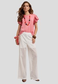 Haut rose à volants à manches courtes, pantalon blanc à jambes larges avec des trous en dentelle, collier superposé, bracelets marron, tenant un sac à main rose.