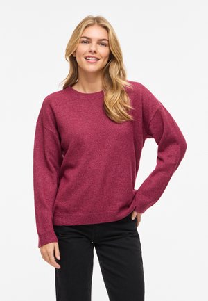 Femme souriante aux longs cheveux blonds, portant un pull à manches longues magenta et un pantalon noir, debout avec une main sur la hanche.