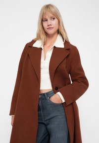 Hnedý kabát z vlneného zmesi s klopovým golierom, veľkými klopami, prednými gombíkmi a vreckami. Nosený cez biely pletený top a modré džíny.