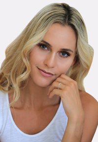 Blond kvinde med bølget hår poserer med hånden på hagen. Hun har en hvid skjorte på og en sølvring. Glat hud, blå øjne, naturlig makeup.