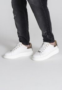 Weiße Sneakers aus glattem Leder mit einem kontrastierenden braunen Akzent an der Ferse, runder Zehenpartie und dicker weißer Sohle.