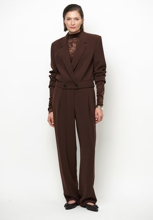 Vrouw draagt een oversized bruin pak met een gestructureerde coltrui-blouse en zwarte puntige platte schoenen, staand tegen een witte achtergrond.