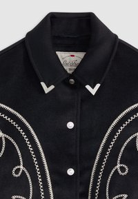 Chemise en laine noire avec boutons-pression argentés, broderie blanche décorative, et pointes de col pointues. Étiquette Levi's visible à l'intérieur.
