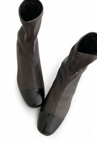 Botas de tobillo marrones de material suave con una parte superior texturizada, punteras en charol negro y costuras decorativas a lo largo del costado.