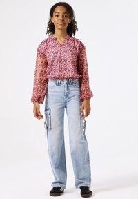 Blouse à motifs rouges et roses avec de longues manches bouffantes et un col en V. Jean bleu clair à jambes larges avec poches cargo. Baskets noires.