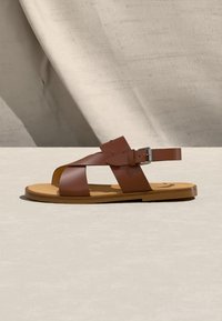 Καφέ δερμάτινο σανδάλι με επίπεδη σόλα, διασταυρωμένους ιμάντες, αγκράφα στον αστράγαλο και ταμπά σόλα πάνω σε ουδέτερο υφασμάτινο φόντο.