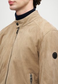 Άνδρας που φοράει μπεζ σουέτ ζακέτα με φερμουάρ και κουμπί στο κολάρο, καθώς και μαύρη στρογγυλή ετικέτα λογότυπου στο επάνω μανίκι, πάνω από ναυτικό μπλούζα με ψηλό λαιμό.