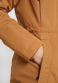 Brązowa kurtka ocieplana o gładkiej teksturze, z mankietami na rękawach i boczną kieszenią dla łatwego dostępu.