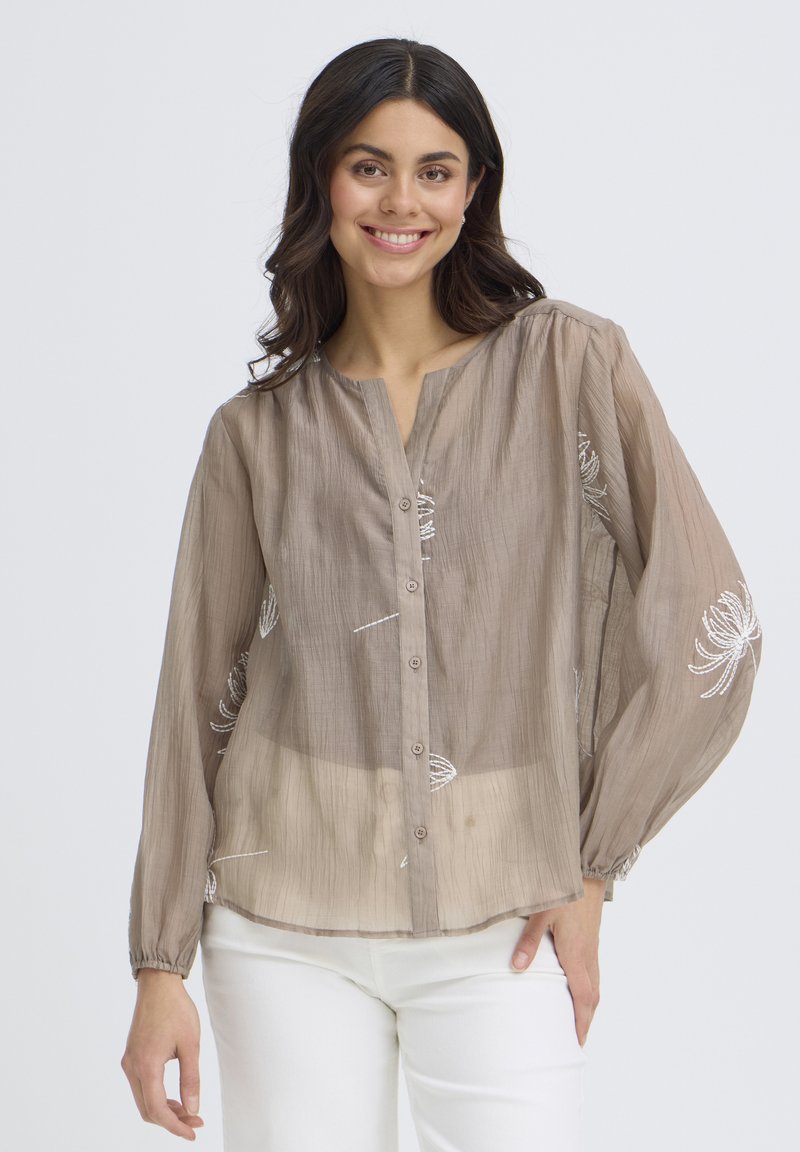Femme souriante portant un chemisier transparent taupe avec des broderies florales blanches et un pantalon blanc, debout avec une main sur la hanche.
