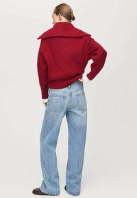 Roter gerippter Pullover mit großem Kragen, kombiniert mit hellblauen Jeans mit weitem Bein. Das Material wirkt weich mit einer lässigen Passform.