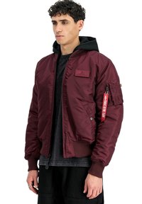 Bordeauxfarbene Bomberjacke aus glänzendem Nylon mit gerippten Bündchen und Saum. Verfügt über eine Fronttasche und ein rotes Etikett am Ärmel.