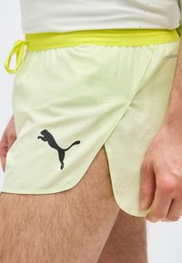 Gros plan sur une personne portant un short Puma vert clair avec une ceinture jaune et une fente latérale, montrant une partie de la jambe et de la main.