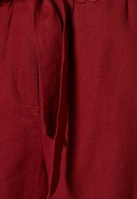 Tejido de lino burdeos con un acabado texturizado; presenta una cintura atada y un detalle de bolsillo lateral, mejorando su diseño funcional.