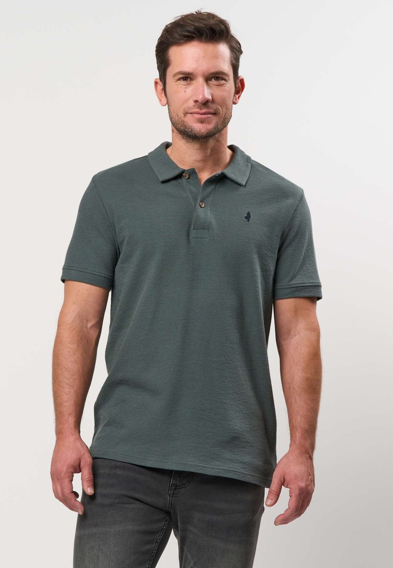 Homme aux cheveux courts et bruns, portant un polo vert foncé à manches courtes et un jean foncé, debout devant un fond clair uni.