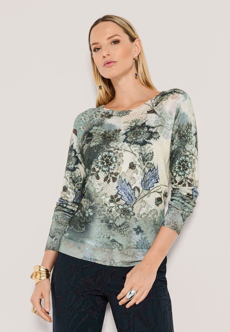 Vrouw in een lange mouwen bloemenpatroon top in groene en blauwe tinten, zwarte gestructureerde broek en gouden en zilveren sieraden.