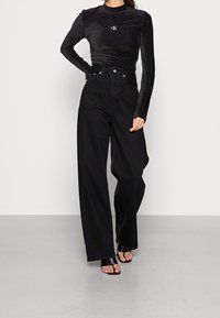 Schwarzes Langarmoberteil aus weichem Stoff mit Logo, kombiniert mit hoch taillierten, weiten schwarzen Jeans. Abgerundet mit schwarzen Slippers.