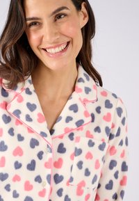 Maglione da pigiama in pile con pattern di cuori multicolori in rosa e blu scuro su uno sfondo crema. Presenta un colletto a punte e una tasca frontale.
