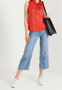 Röd ärmlös topp med vita prickar och en rosettdetalj, tillsammans med stålliga blå jeans och vita sneakers, bärande på en svart väska.