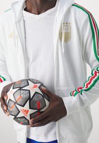 Veste de sport blanche zippée avec des rayures vertes, rouges et blanches, emblème doré de l'Italie, accompagnée d'un ballon de football noir et blanc à motif.
