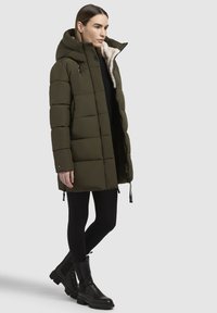 Geacă căptușită de culoare verde olive, cu glugă, având o căptușeală din blană sintetică, cu fermoar în față și șnururi ajustabile la tiv și manșete.