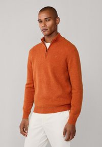 Felpa arancione con zip, in maglia testurizzata, maniche lunghe e orlo a coste. Indossata sopra una camicia bianca, abbinata a pantaloni chiari.
