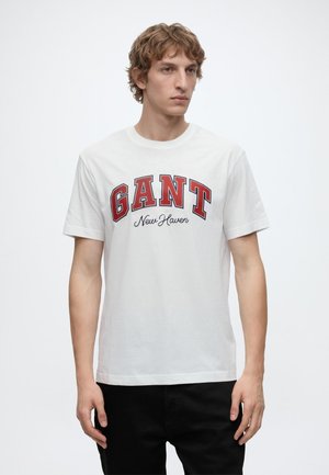 Junger Mann mit lockigem Haar, der ein weißes T-Shirt mit rotem "GANT" und schwarzem "New Haven" Text trägt, kombiniert mit schwarzen Hosen, steht vor weißem Hintergrund.