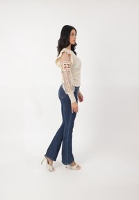 Maglione beige a coste con spalle arricciate e maniche lunghe trasparenti e stampate. Jeans a zampa d'elefante blu scuro e décolleté bianchi con apertura sulla punta e design testurizzato.