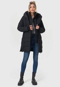Schwarze Pufferjacke mit Kapuze, gestepptem Design, Reißverschluss vorne und Knopfverschlüssen. Getragen über einem grauen Oberteil und kombiniert mit blauen Jeans und schwarzen Stiefeln.
