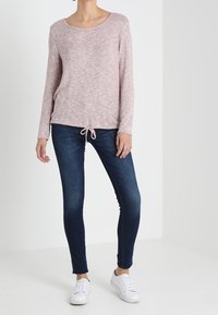 Maglietta a maniche lunghe rosa chiaro con scollatura a barca e orlo regolabile abbinata a jeans slim-fit blu scuro e sneakers bianche.