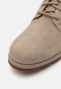 Pantofle din suede bej cu vârf rotunjit, superior texturat și talpă din cauciuc contrastantă. Prezintă șireturi plate și cusături pe mijlocul tălpii.