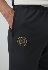 Main reposant dans la poche d'un pantalon de sport noir avec un écusson du logo du Paris Saint-Germain en or et noir sur la cuisse.