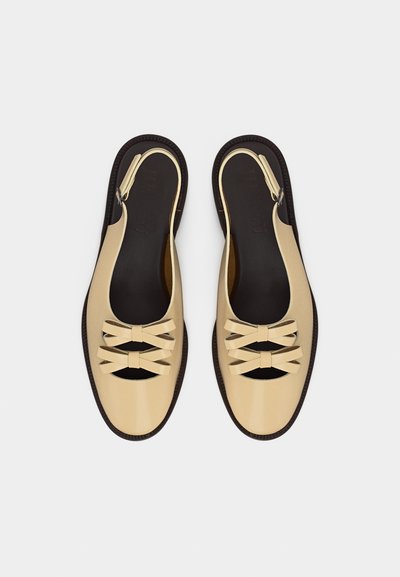 Μπεζ επίπεδα παπούτσια slingback με σκούρες σόλες και διπλές φιόγκους στο μπροστινό μέρος, τοποθετημένα δίπλα-δίπλα σε λευκό φόντο.