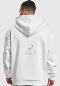 Witte hoodie met een capuchon en een achterkant met een grafisch ontwerp van paarse veren en tekst. Gemaakt van zachte, textuurrijke stof met geribbelde manchetten.