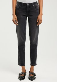 Žena oblečená v černých úzkých džínách s vysokým pasem a černých špičatých botách s několika přezkami a kovovými oky.