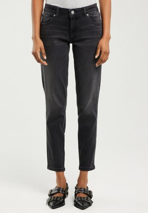 Žena oblečená v černých úzkých džínách s vysokým pasem a černých špičatých botách s několika přezkami a kovovými oky.