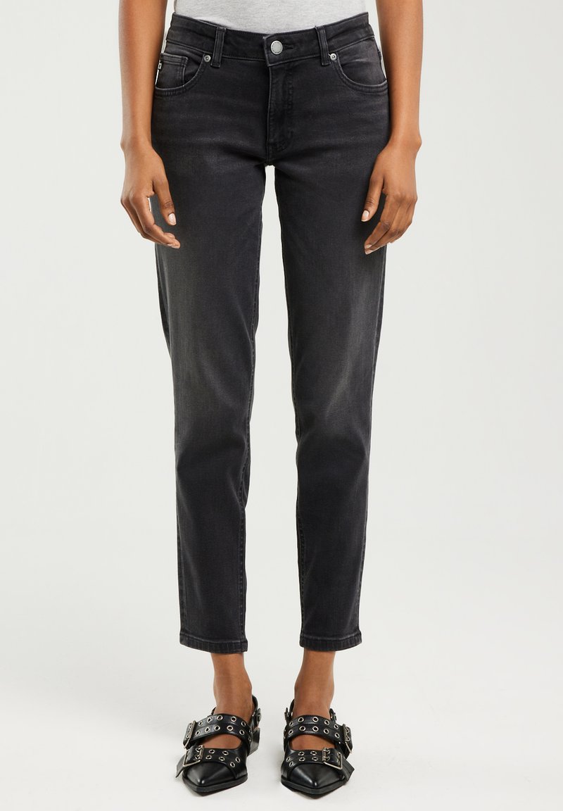 Žena oblečená v černých úzkých džínách s vysokým pasem a černých špičatých botách s několika přezkami a kovovými oky.