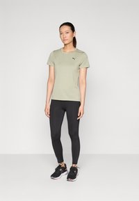 Olivgrön träningströja med kort ärm och svart logotyp, tillsammans med svarta leggings och svarta träningsskor med rosa detaljer.