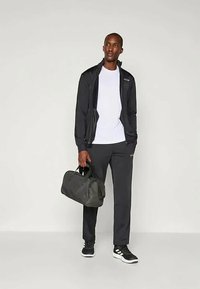 Crna sportska jakna sa zatvaračem, bijela majica, crne trenirke i crne tenisice. Drži tamnu torbu. Jednostavan dizajn, funkcionalan stil.