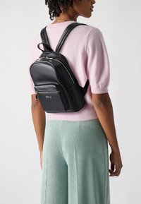 Zaino in pelle nera con scomparti con zip, spallacci regolabili e una piccola tasca frontale. Indossato sopra un maglione rosa chiaro e pantaloni verdi.
