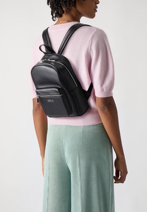 Black leather backpack with zippered compartments, adjustable straps, and a small front pocket. Worn over a light pink sweater and green pants.