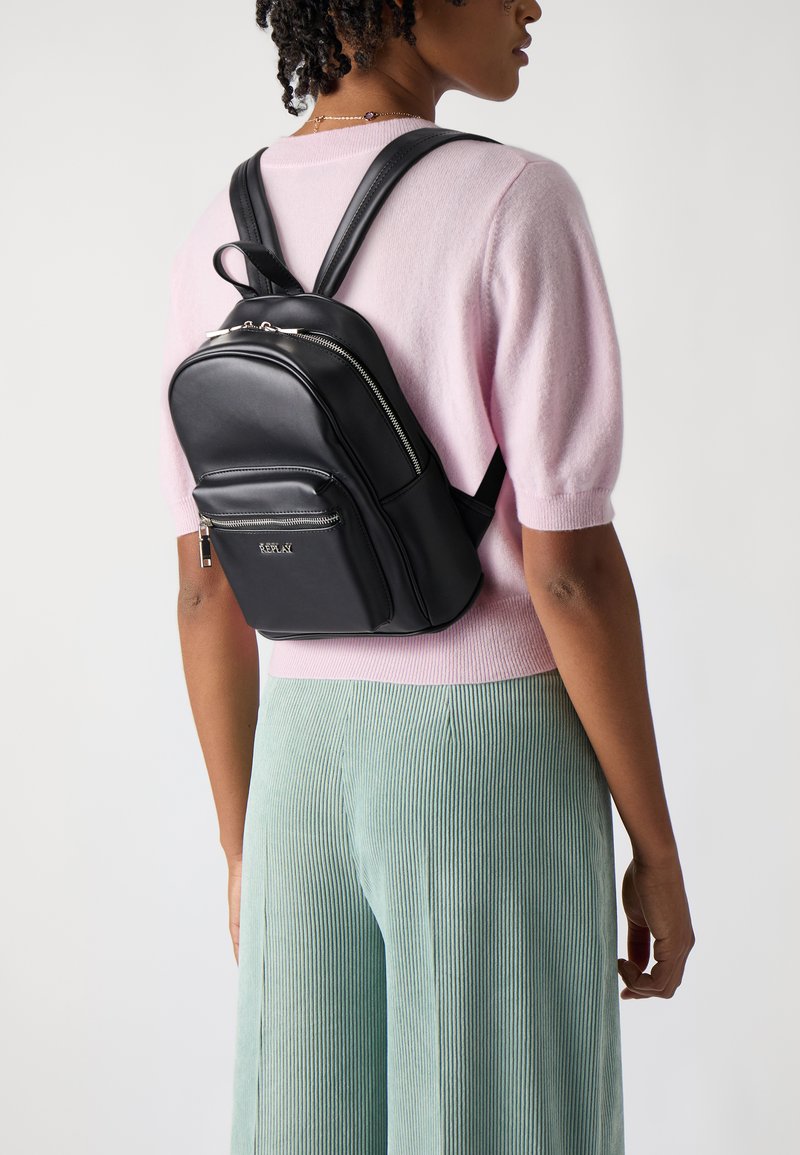 Zaino in pelle nera con scomparti con zip, spallacci regolabili e una piccola tasca frontale. Indossato sopra un maglione rosa chiaro e pantaloni verdi.