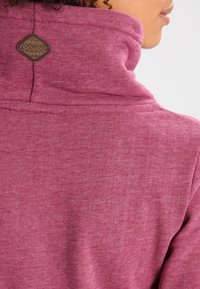 Dettaglio di una persona che indossa una felpa con colletto alto color bordeaux, mostrando il retro con una piccola toppa decorativa marrone vicino al colletto.
