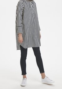 Túnica larga a rayas en azul marino y blanco, con cuello abotonado, mangas anchas y aberturas laterales. Combinada con jeans ajustados oscuros y zapatillas blancas.