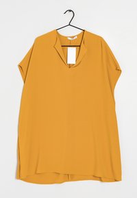 Blusa mostaza de manga corta con escote en forma de V y aberturas laterales. Hecha de una tela suave, con una etiqueta adjunta en el cuello.