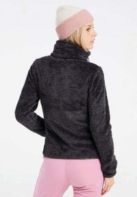 Donkergrijze fleecejack met hoge kraag, voorzien van een gestructureerd oppervlak. Draagt een roze en witte muts. Roze yogabroek zichtbaar eronder.