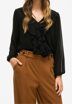 Kvinde iført en sort gennemsigtig bluse med lange ærmer og flæser samt højtsiddende brune bukser med knapdetaljer, hænderne i lommerne.