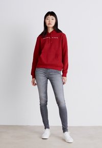Červená bavlnená mikina s kapucňou a predným vreckom, spojená so šedými slim-fit džínsami a bielymi teniskami. Jednoduchý, neformálny dizajn.