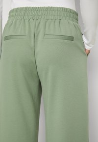 Un pantalon de survêtement vert avec une taille élastique à cordon, fabriqué en tissu doux, comportant deux poches arrière en patch et une texture lisse.