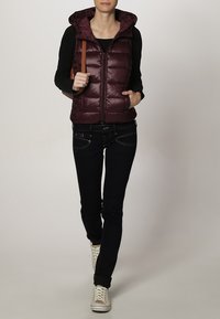 Bordeauxrote Steppweste mit Kapuze und vertikalem Steppmuster, kombiniert mit schwarzer Jeans und weißen Sneakern, mit einem beigen Schulterriemen.